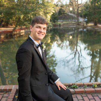 A young man in a tuxedo sitting on the side of a pond.